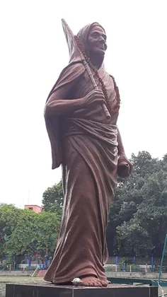 A tall terracotta-coloured statue of Matangini Hazra striding forward with a flagstaff, garlanded, standing outdoors.